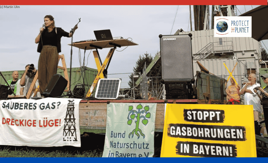 Foto von einer Demonstration gegen Erdgasbohrung am Ammersee, man sieht die Klimaaktivistin Luisa Neubauer auf einer Bühne auf einem Feld am Mikrofon, umgeben von Demonstrations-Schildern und Lautsprechern.